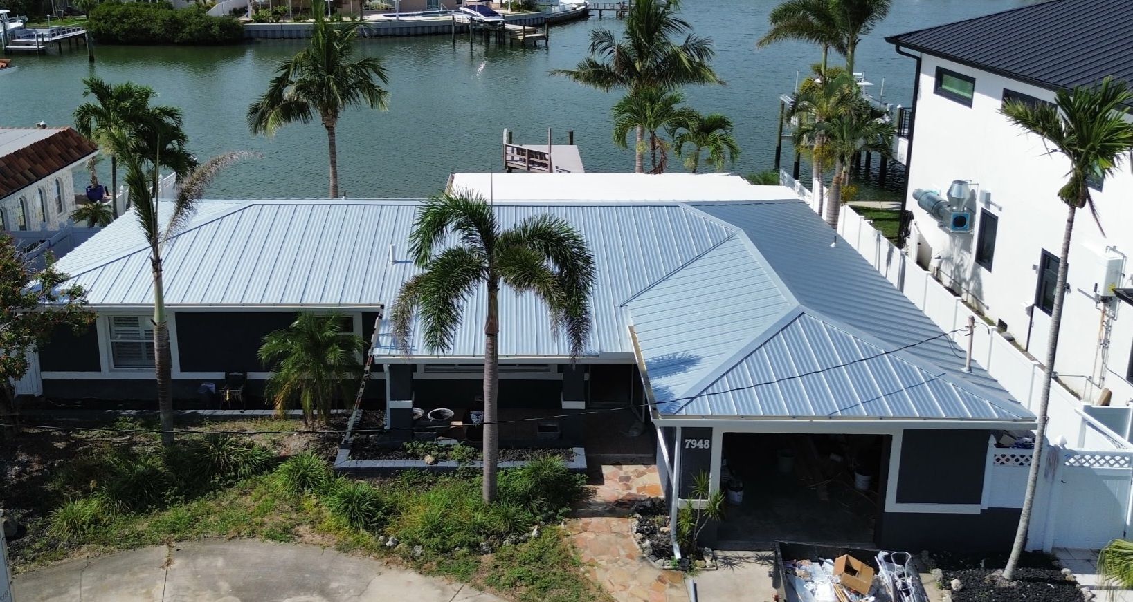 Waterfront home with gray metal roof and garage. Palm trees and boats in the background.