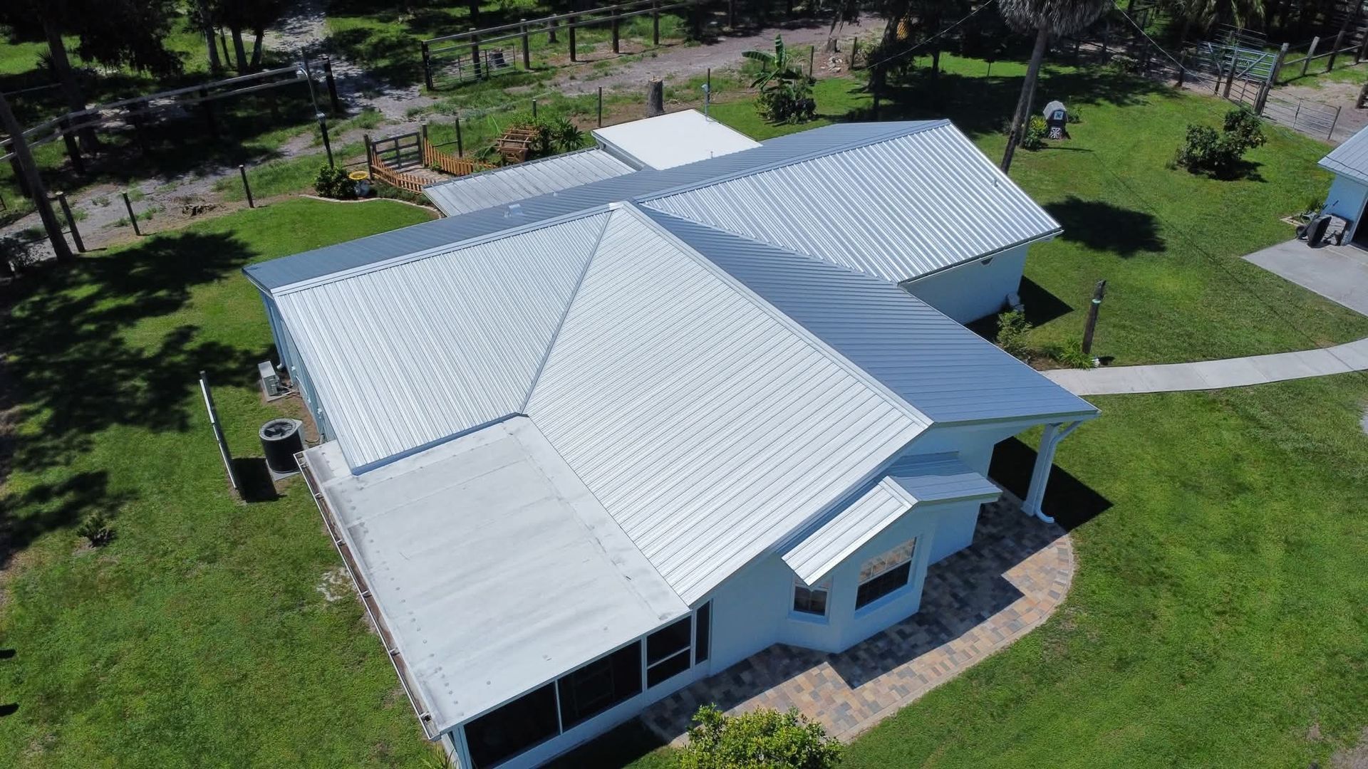 Aerial view of a light blue house with a white metal roof, surrounded by green grass.
