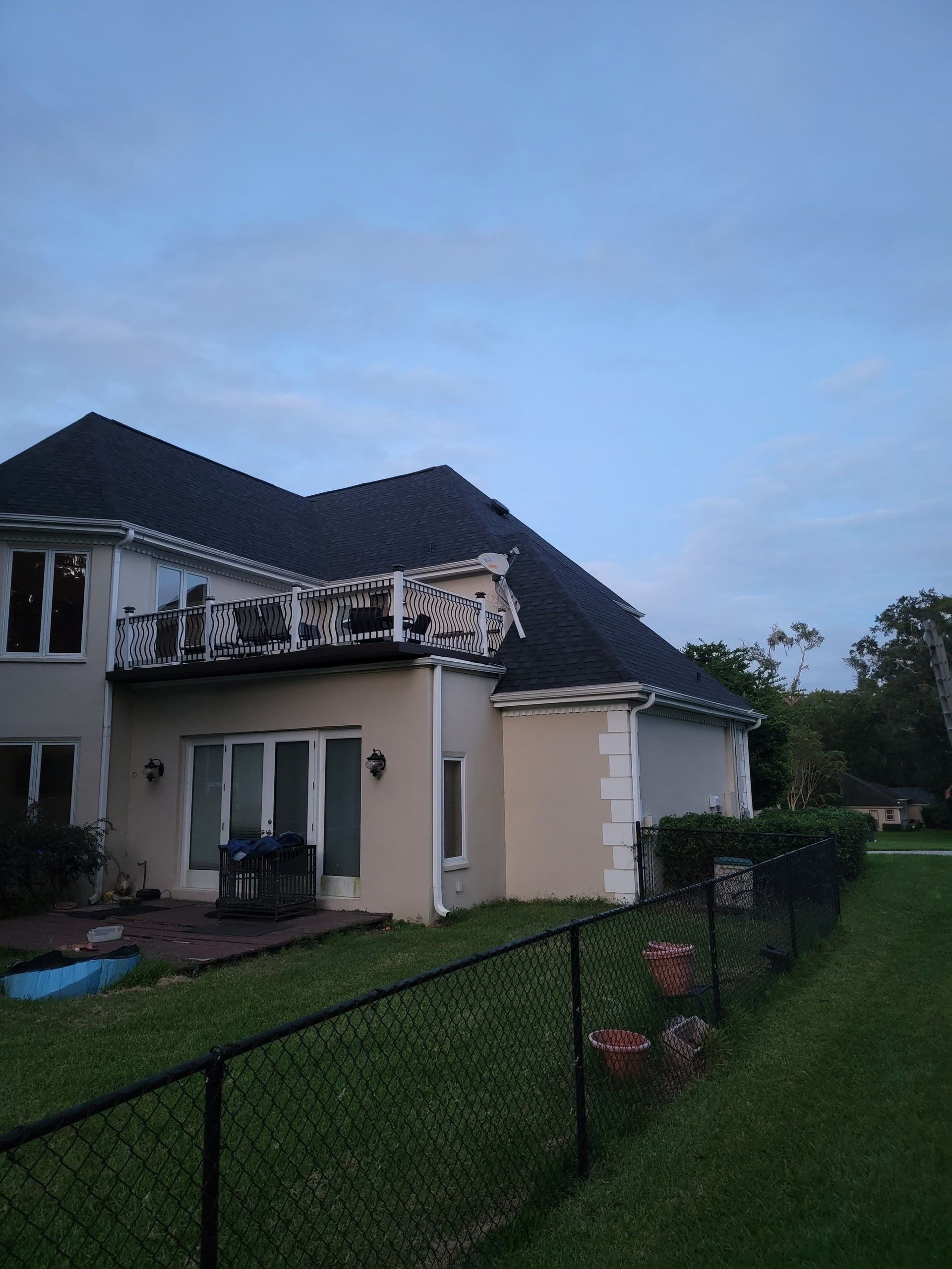 Rear view of a two-story house with a black roof and a balcony, next to a grassy yard and a black fence.