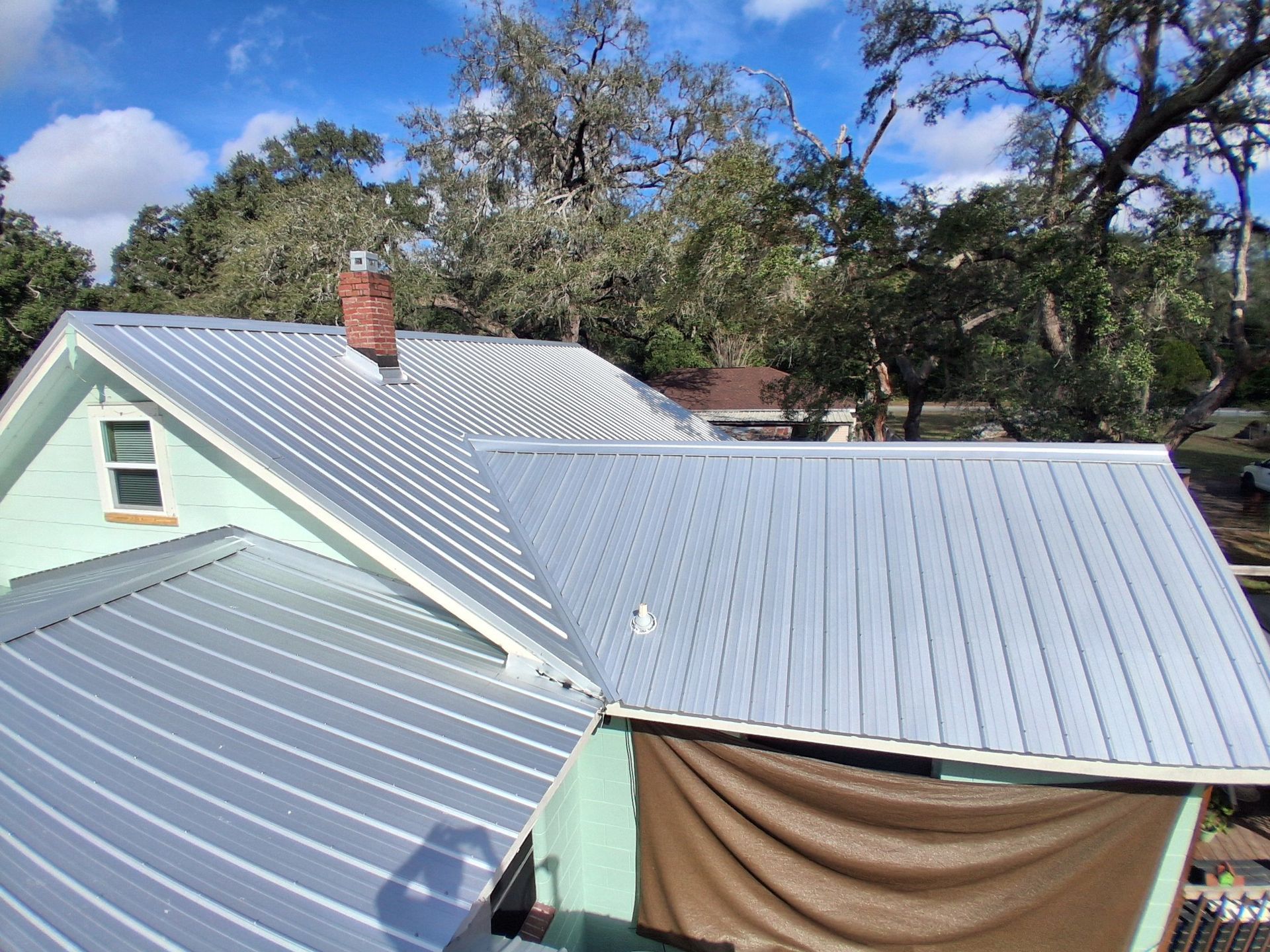 A metal roof on a light green house with a brick chimney under a blue sky.