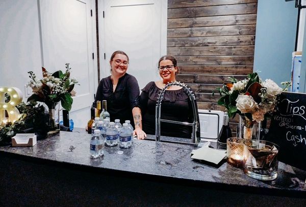 Two women are standing behind a counter with bottles of water and flowers.
