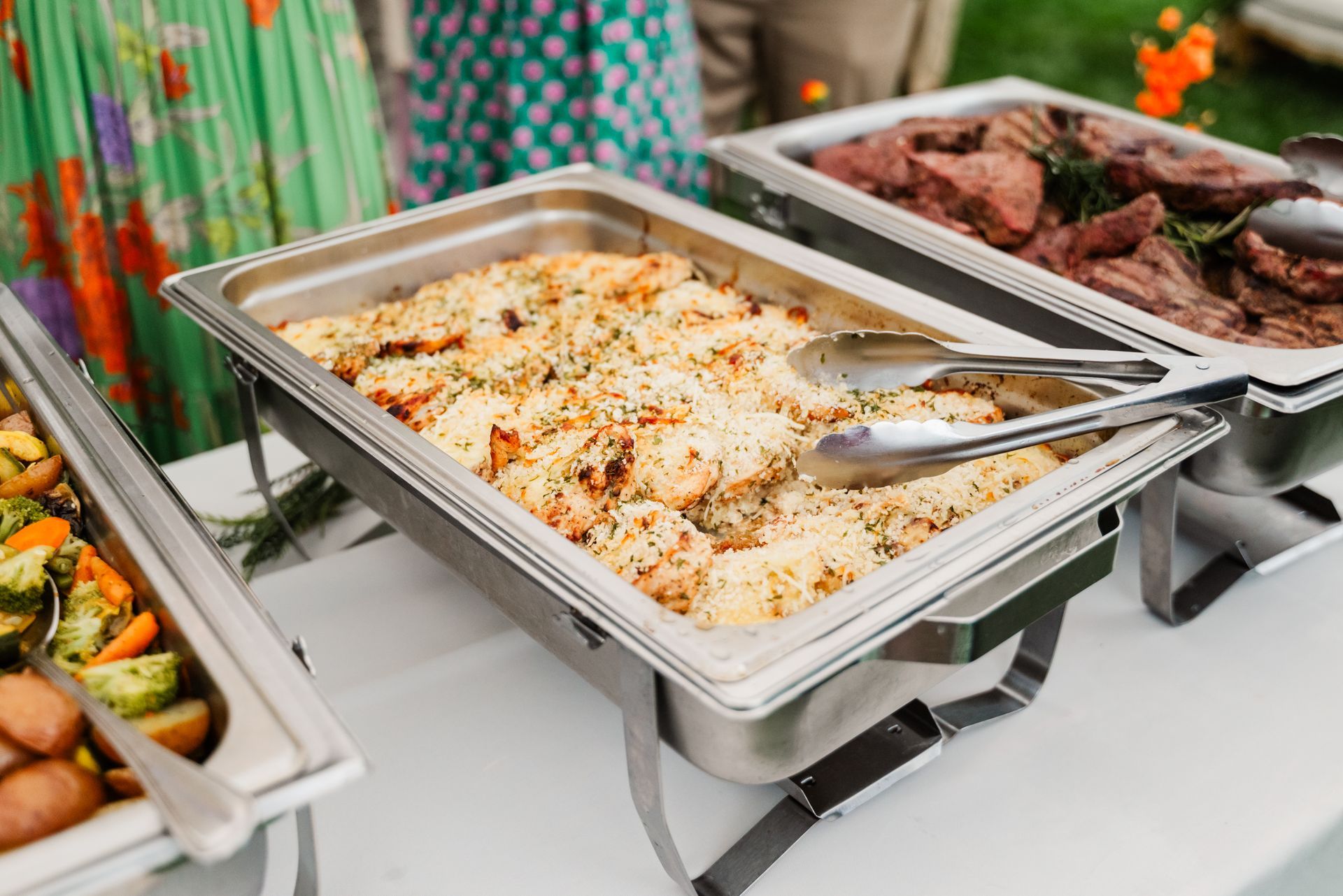 A buffet table with trays of food and tongs on it.
