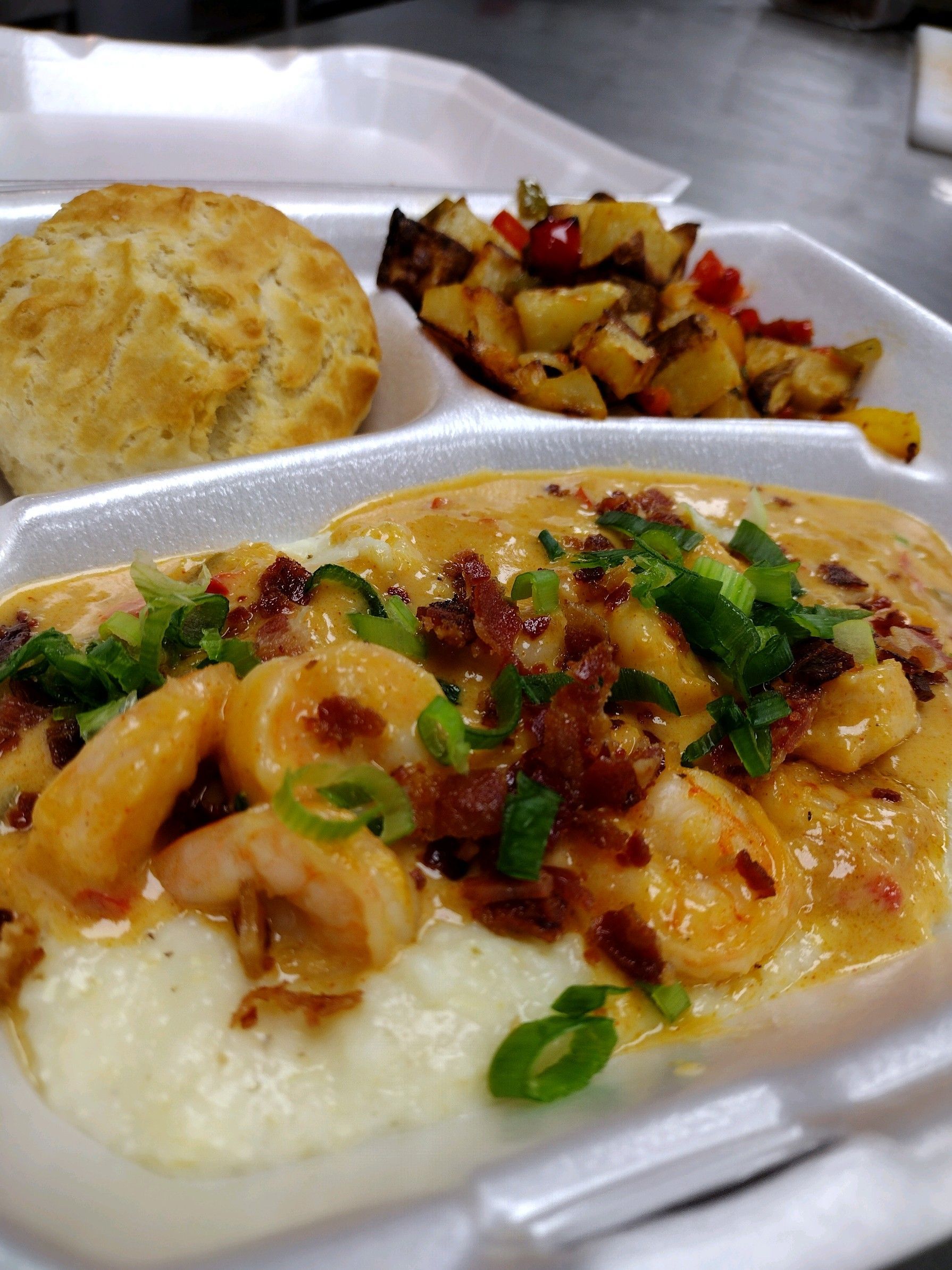 A styrofoam container filled with food including shrimp grits and a biscuit.