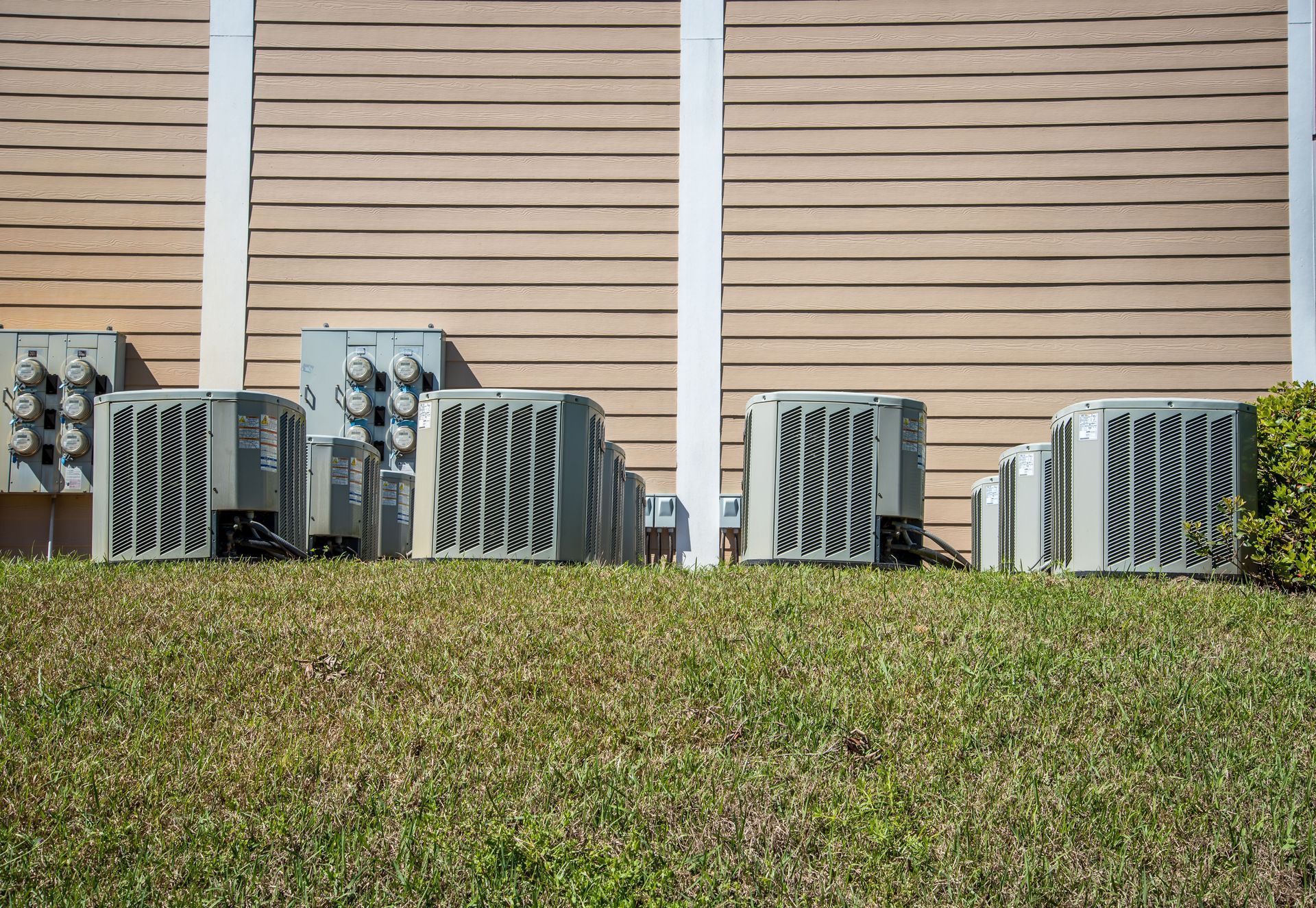 Air conditioning units lined up on a grassy area in front of a building with horizontal siding.