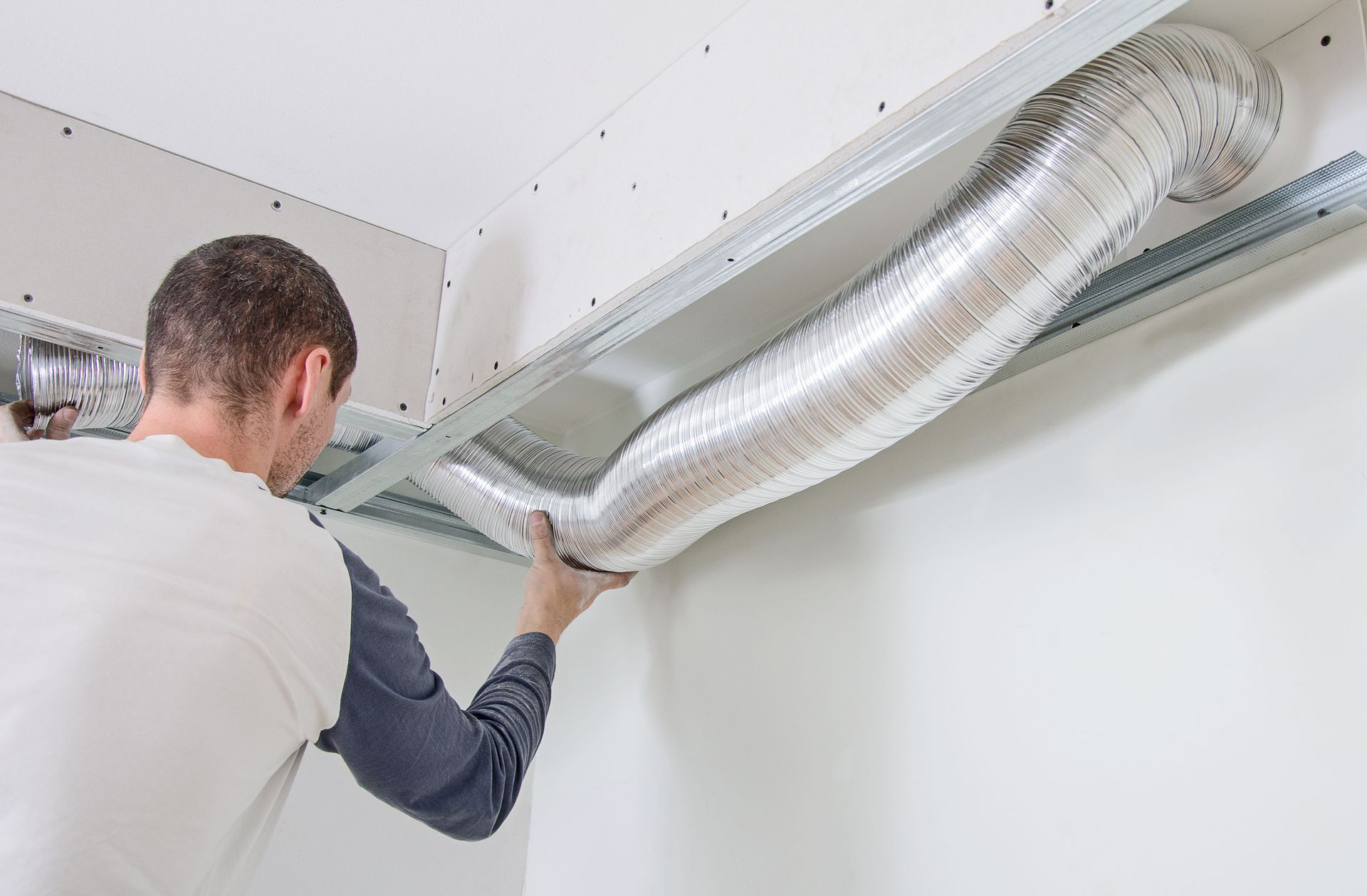 A person installs flexible ductwork within a ceiling, holding the metal tube in place.