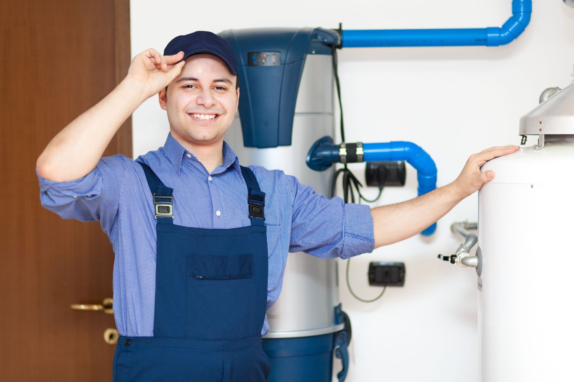 Smiling person in work overalls adjusting cap, standing near water heater and pipes.