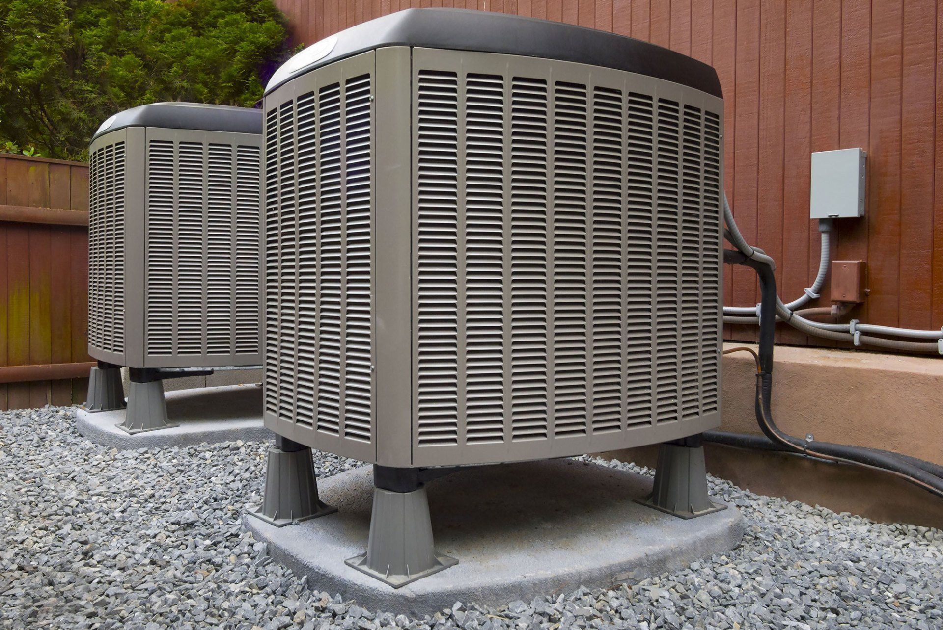 Two air conditioning units on concrete pads, surrounded by gravel, near a wooden fence.
