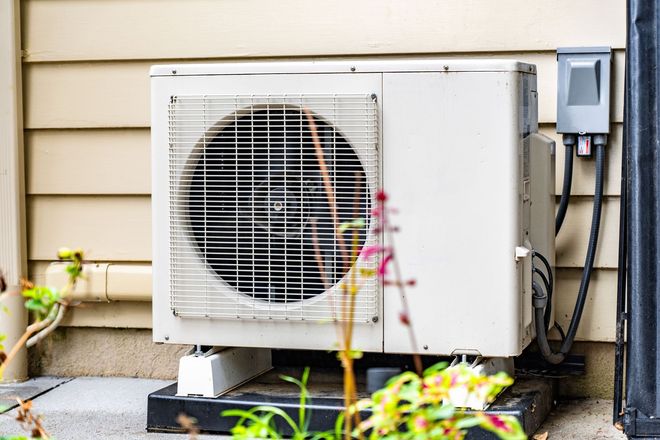 Air conditioning unit mounted on a black base, near a house wall and electrical box, with some plants in the foreground.