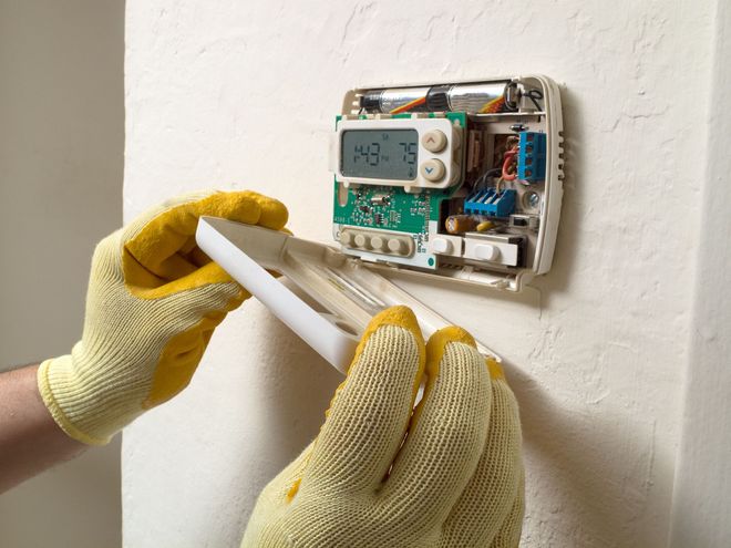 Person in yellow gloves opening a thermostat mounted on a white wall.