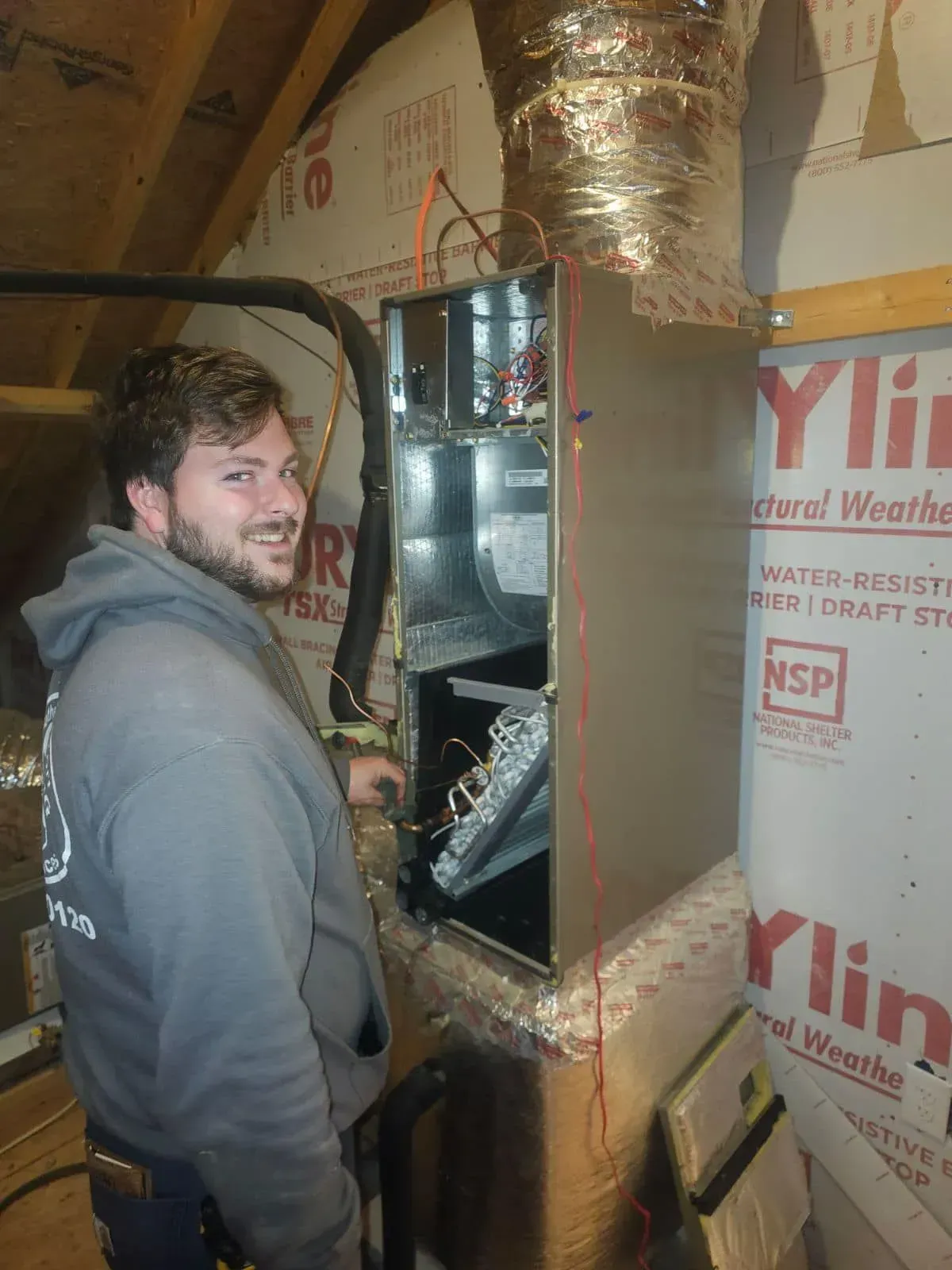 A man smiling next to an HVAC unit in an attic. He's wearing a grey hoodie. The unit has ductwork.