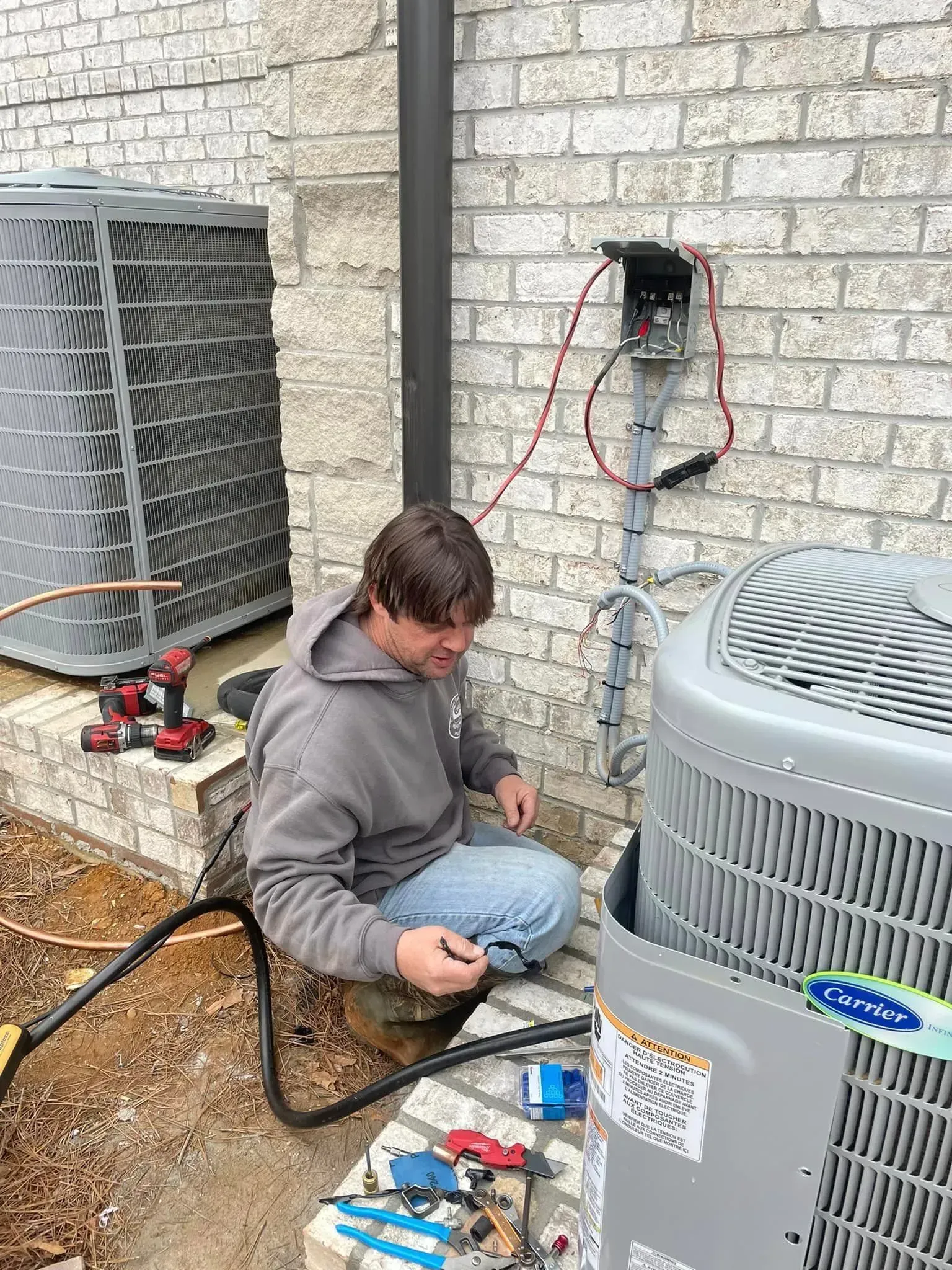 A person works on an HVAC unit next to a brick wall. He is crouching, with tools around him.