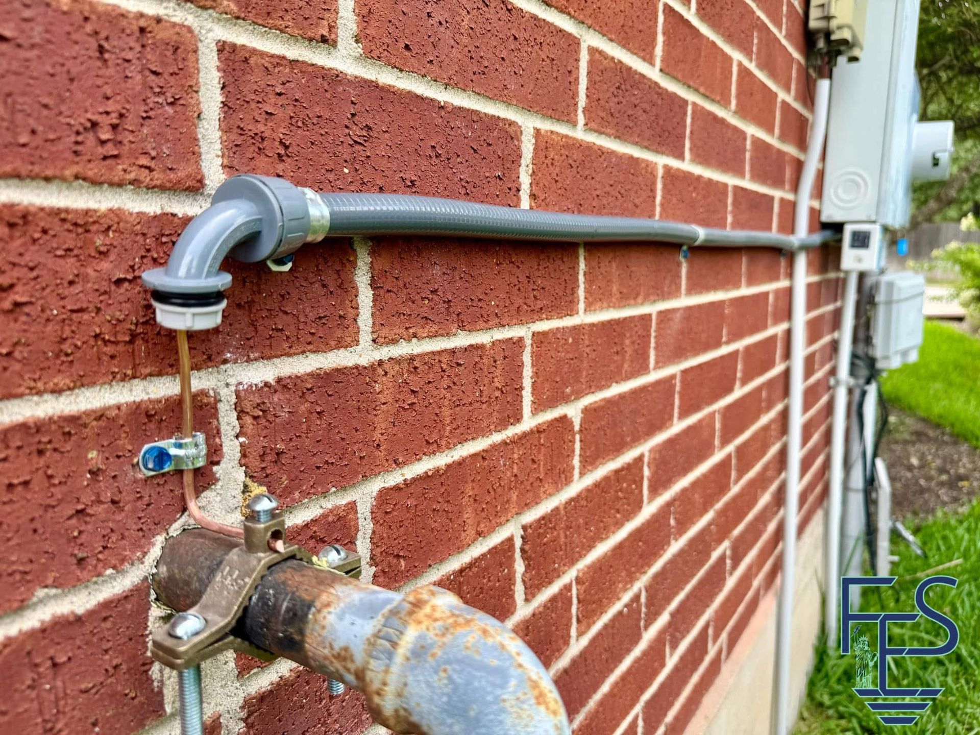 Electrical conduit and grounding wire attached to a rusty pipe on a red brick wall.