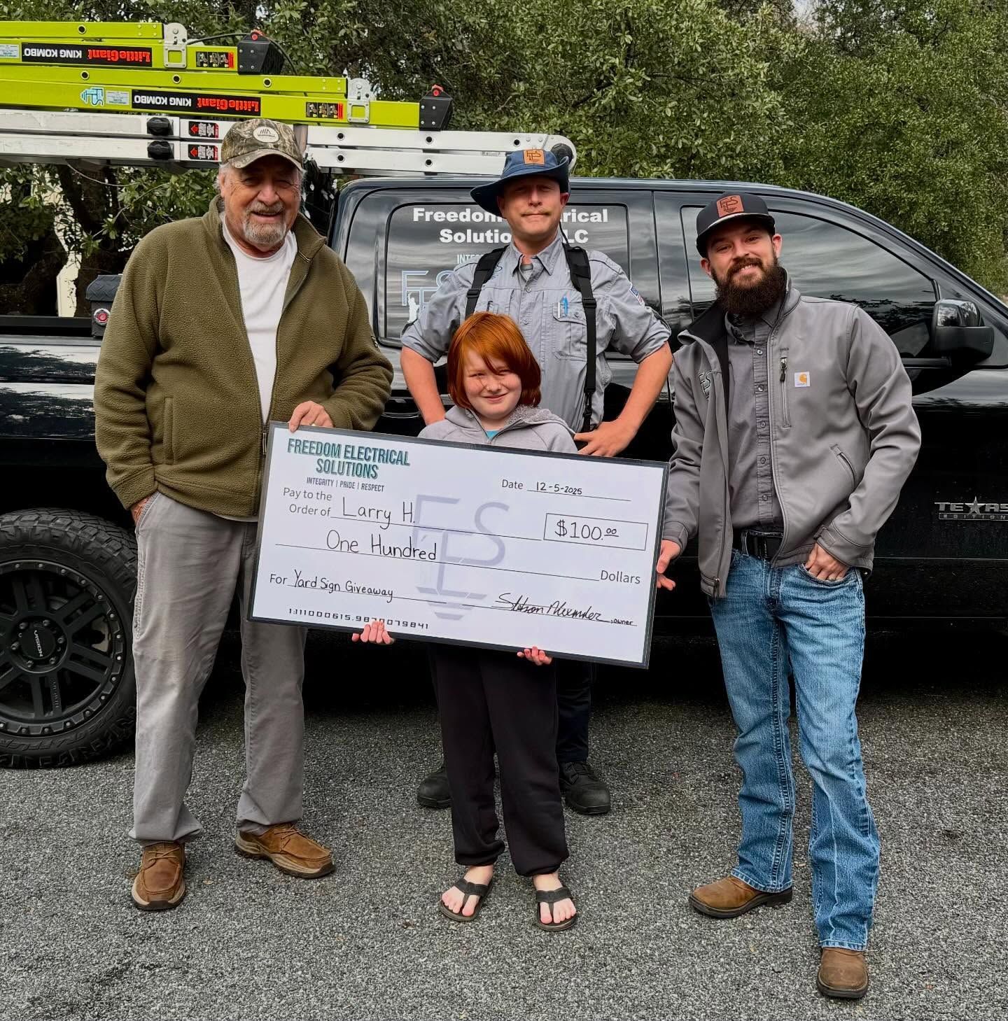 Four people standing near a black truck, holding a large check with logo.