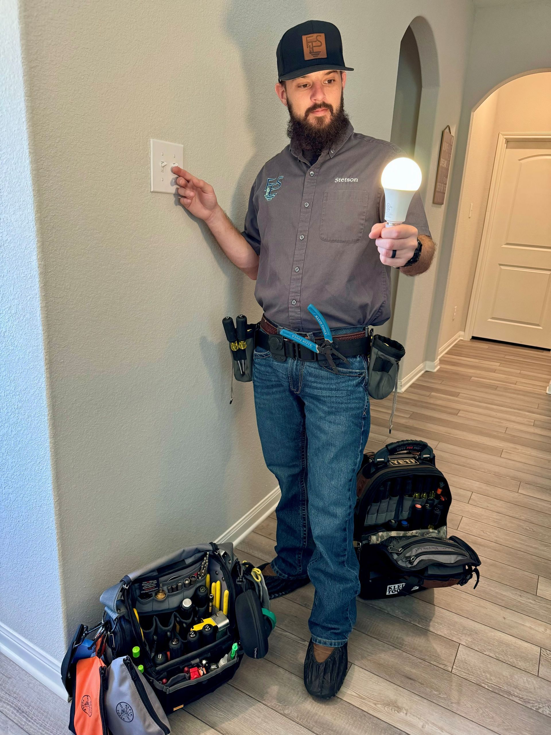 Electrician holding lit lightbulb near light switch; tools at feet.