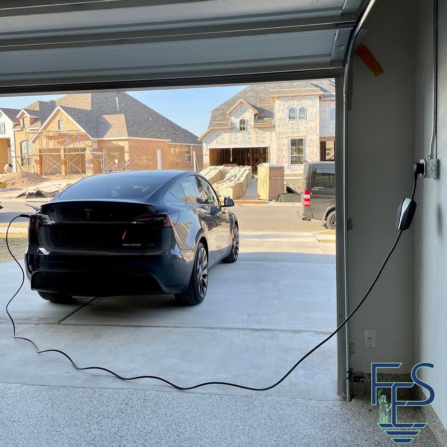 Black Tesla car charging in a garage with houses in the background.