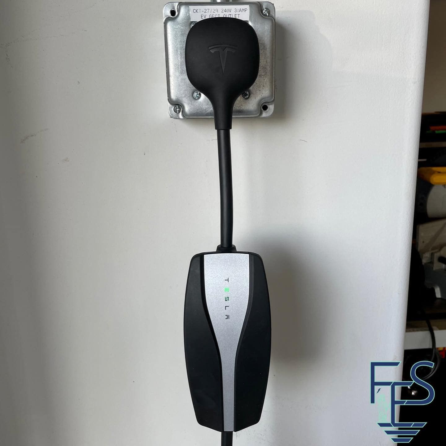 Tesla charging cable plugged into a wall outlet on a white wall.