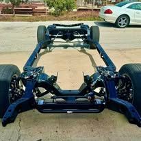 A car chassis is parked on the side of the road next to a white car.