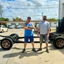 Two men are standing next to a car chassis in a parking lot.
