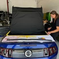 A man is sitting in front of a blue mustang with the hood open.