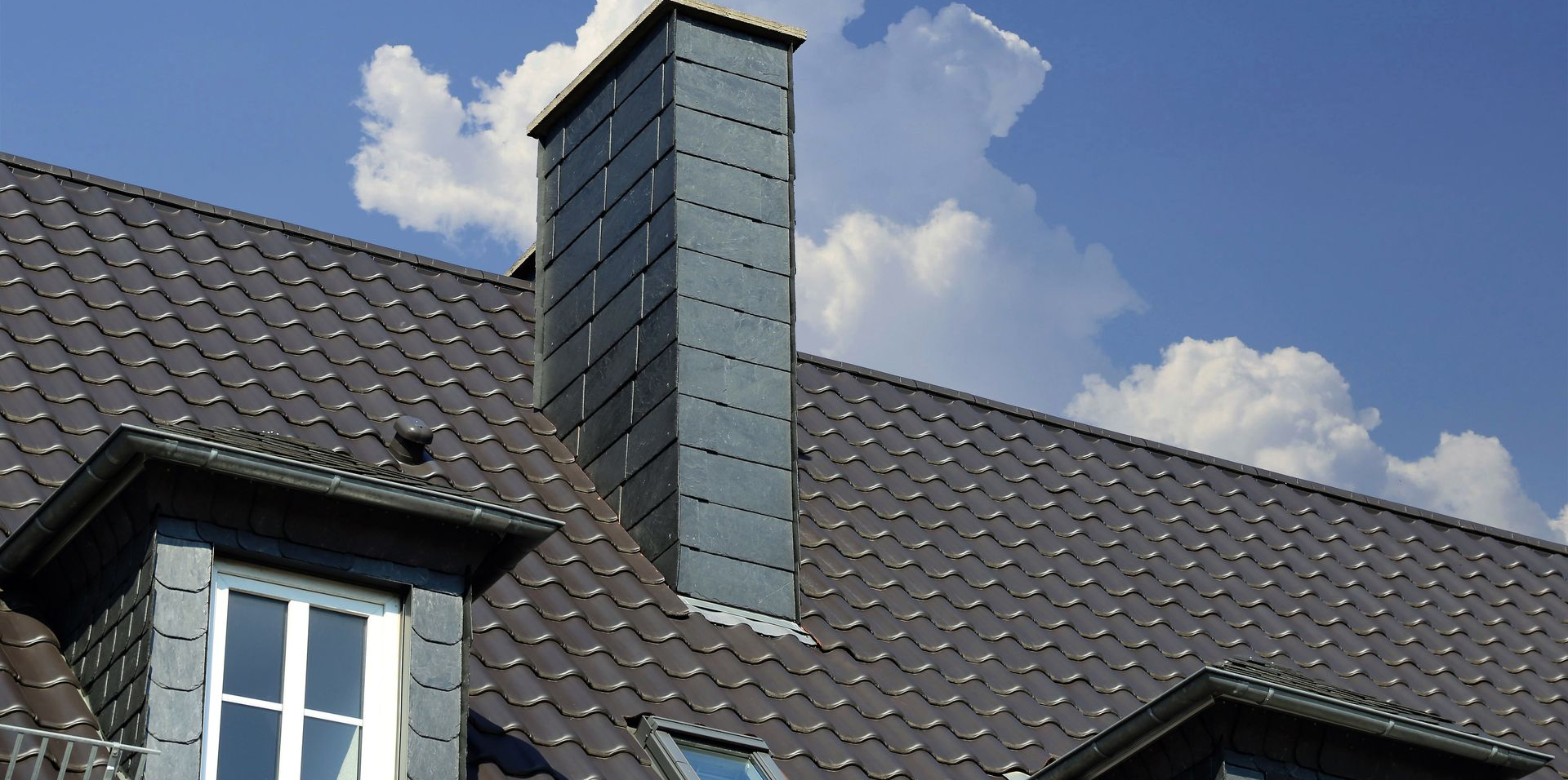 Black tiled roof with a gray brick chimney against a blue sky with clouds