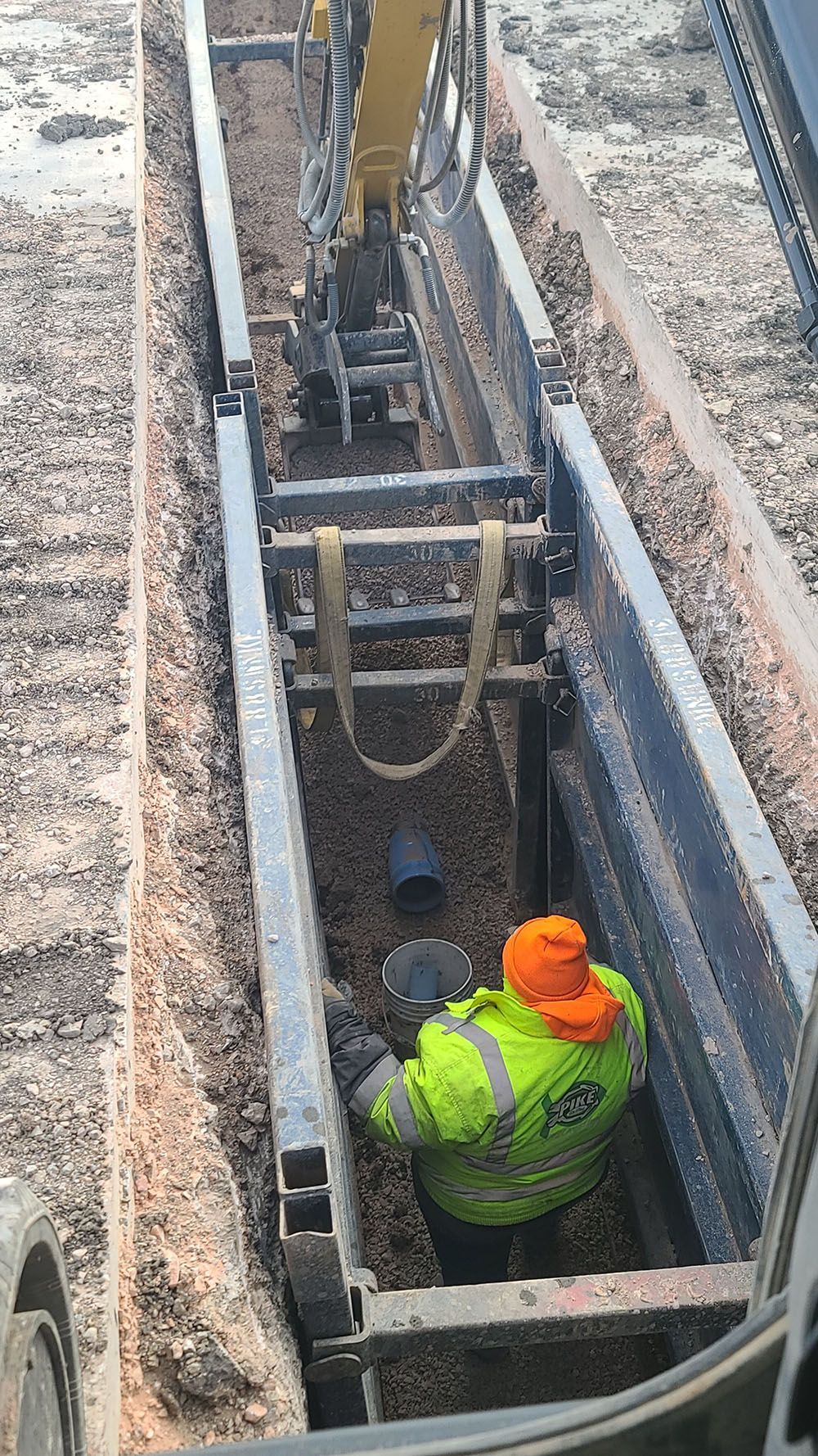 Two workers in safety vests and hard hats inspecting a narrow trench with ladders and pipes