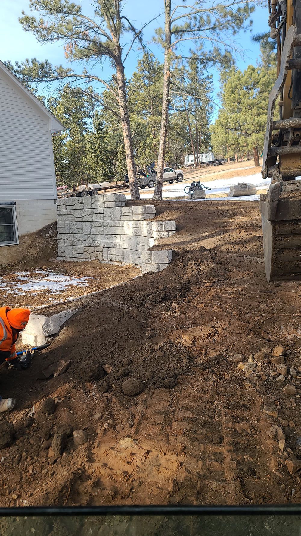 Backyard construction site with dirt mound, retaining wall, trees, and a worker in orange at left