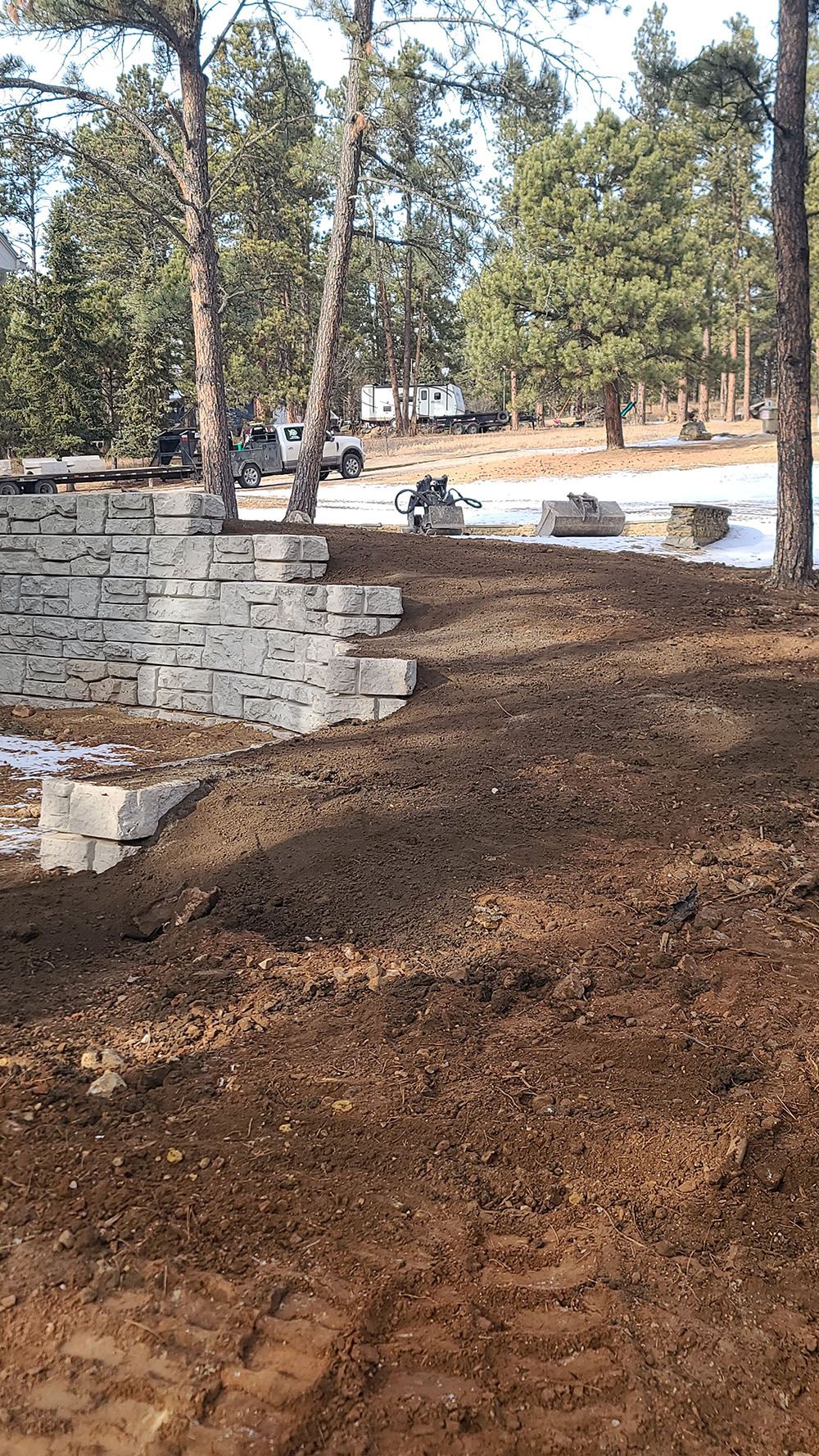 Dirt path beside a low stone wall in a wooded park, with parked cars in the background.