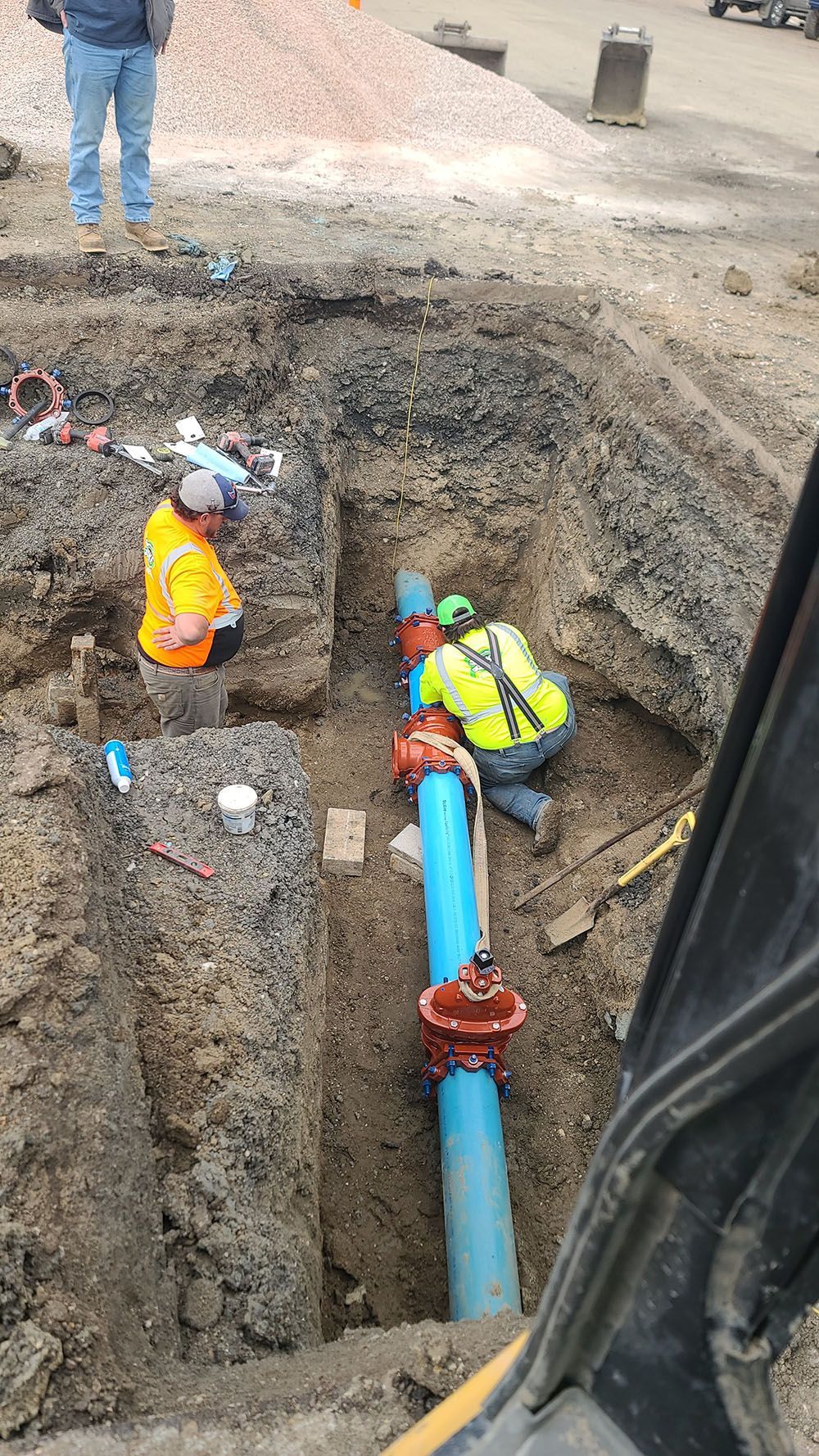 Workers in a trench repairing a blue underground pipe at a construction site.