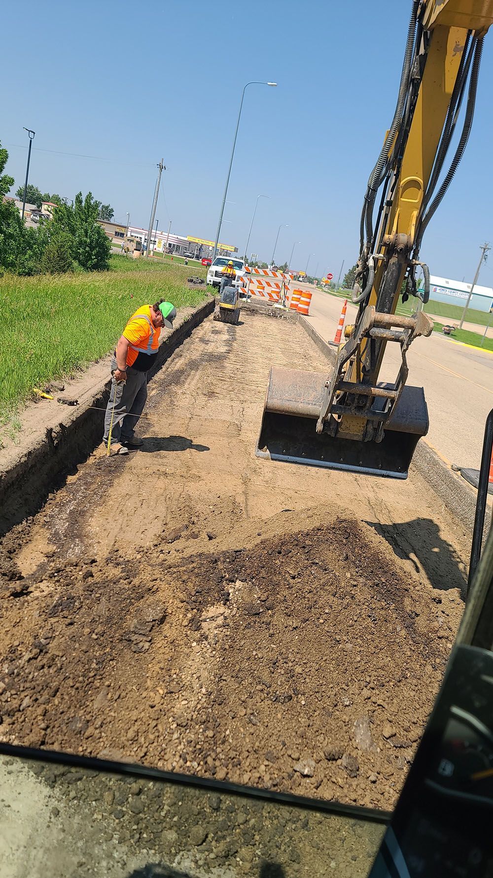 Roadwork scene with excavator loading gravel as a worker in orange vest stands beside a trench.