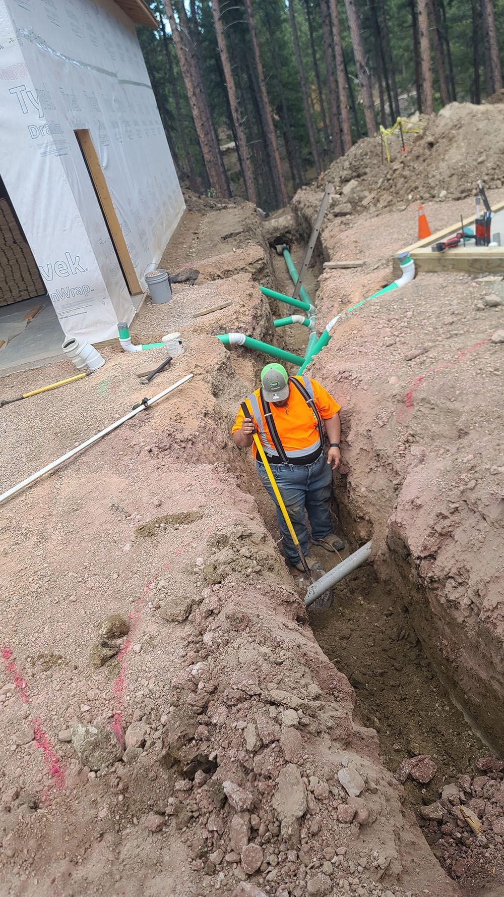 Worker in orange vest standing in a deep trench at a construction site near a house and forest.