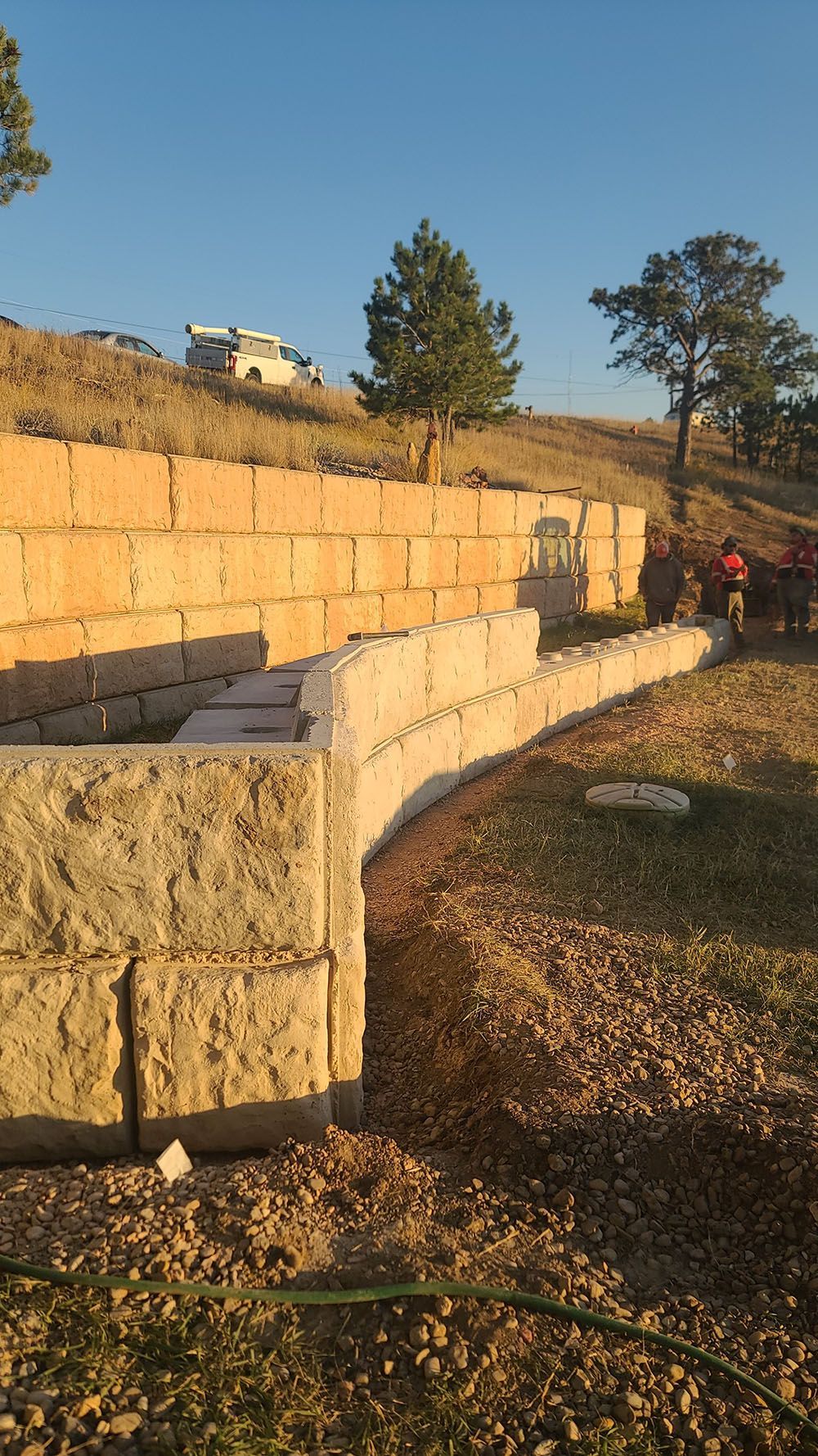 Stone retaining wall along a dirt path at sunset, with trees and a grassy hillside in the background