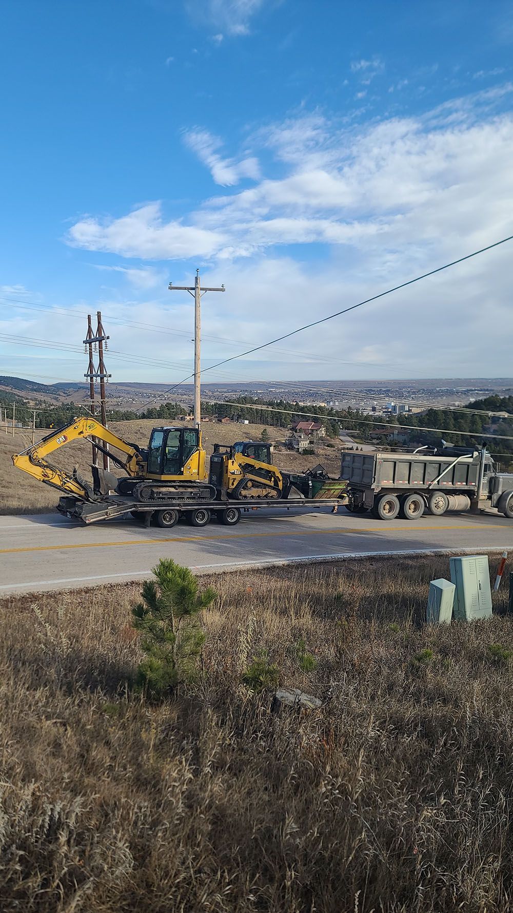 Yellow excavator loading debris onto a trailer on a roadside under a blue sky.