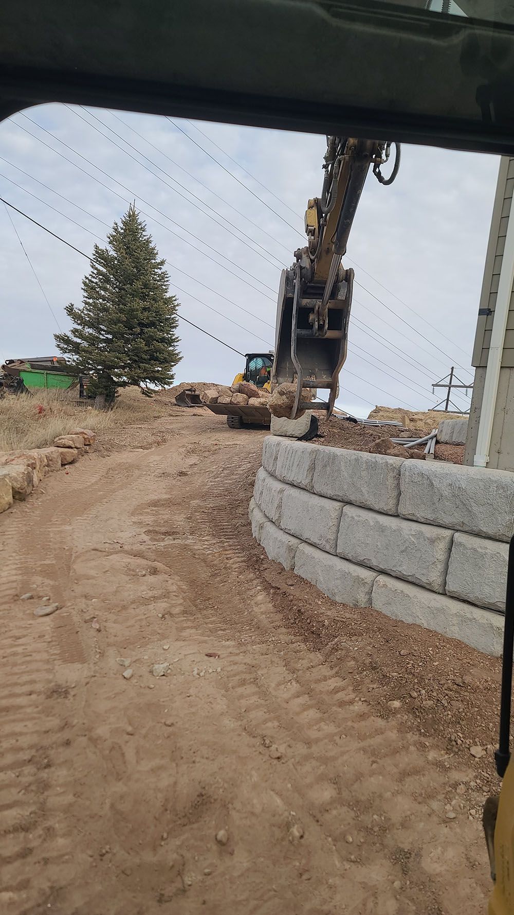 Construction site with excavator beside a dirt road and stacked concrete blocks near a lone tree