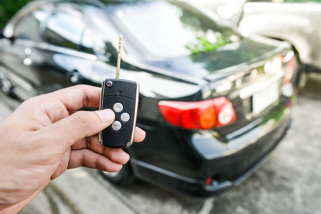 Hand holding car key fob near a black sedan with red taillight