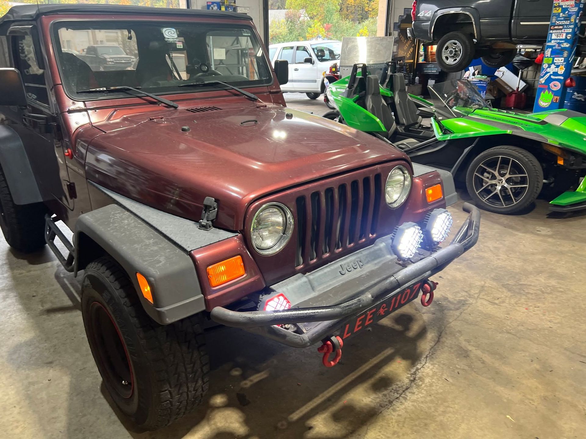 Maroon Jeep Wrangler parked indoors with bright off-road lights and a green buggy in the background