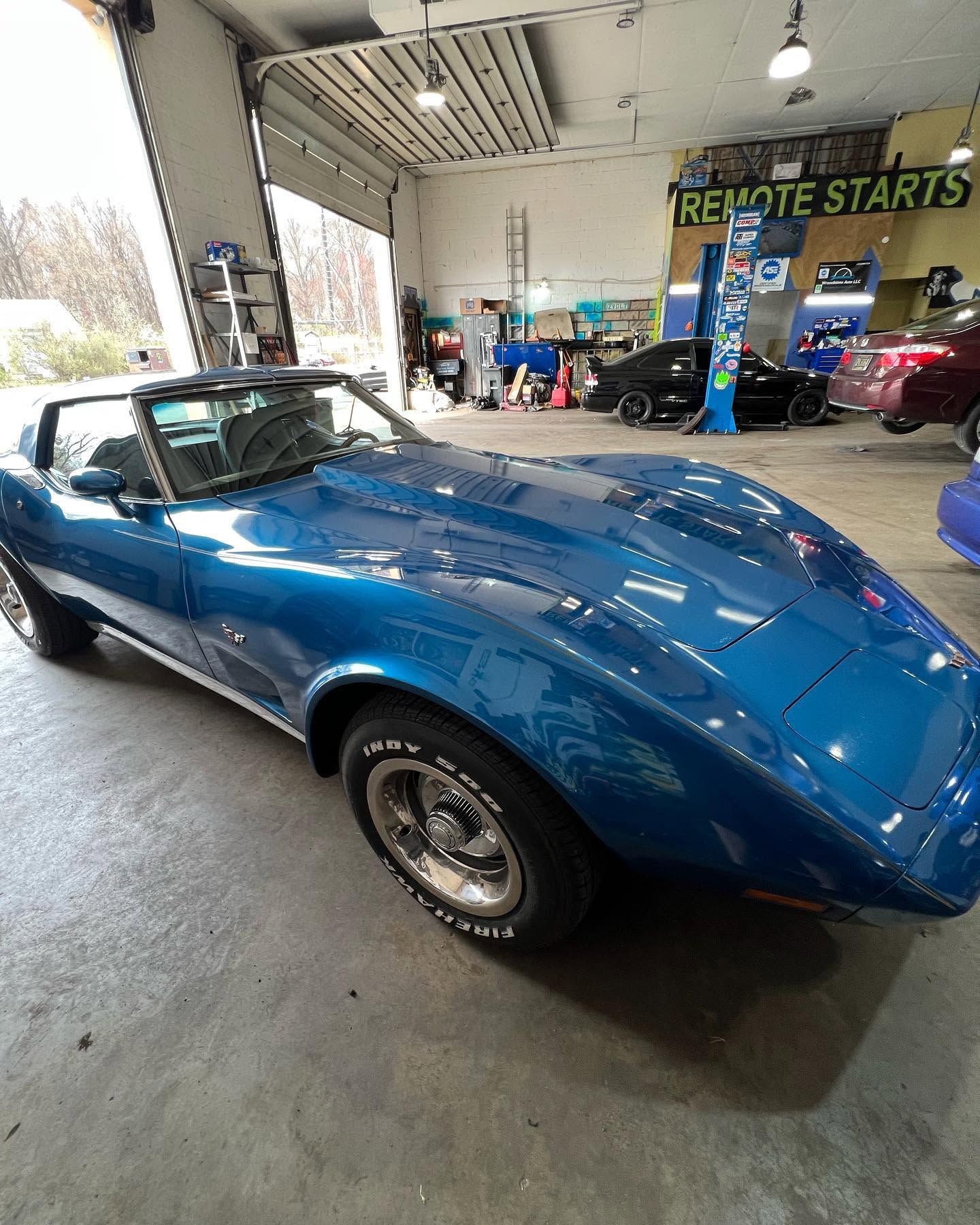Blue classic sports car in an auto shop garage, front-left view under bright lights.