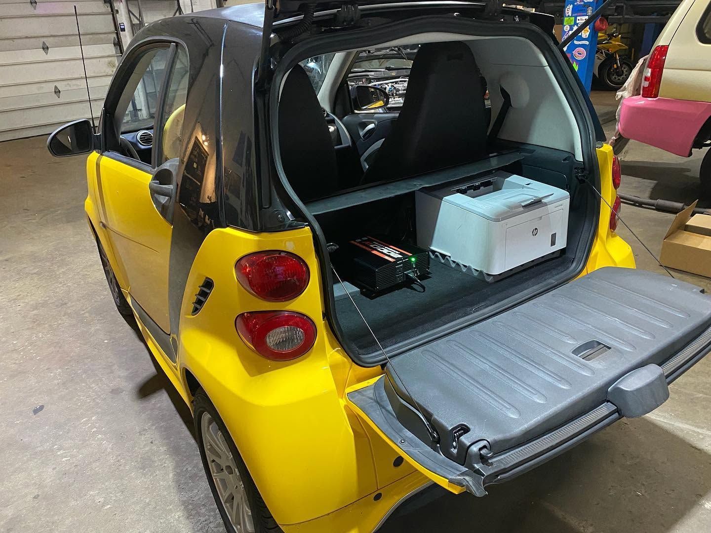 Yellow Smart car with open rear hatch, showing cargo area and a white box inside in a garage.
