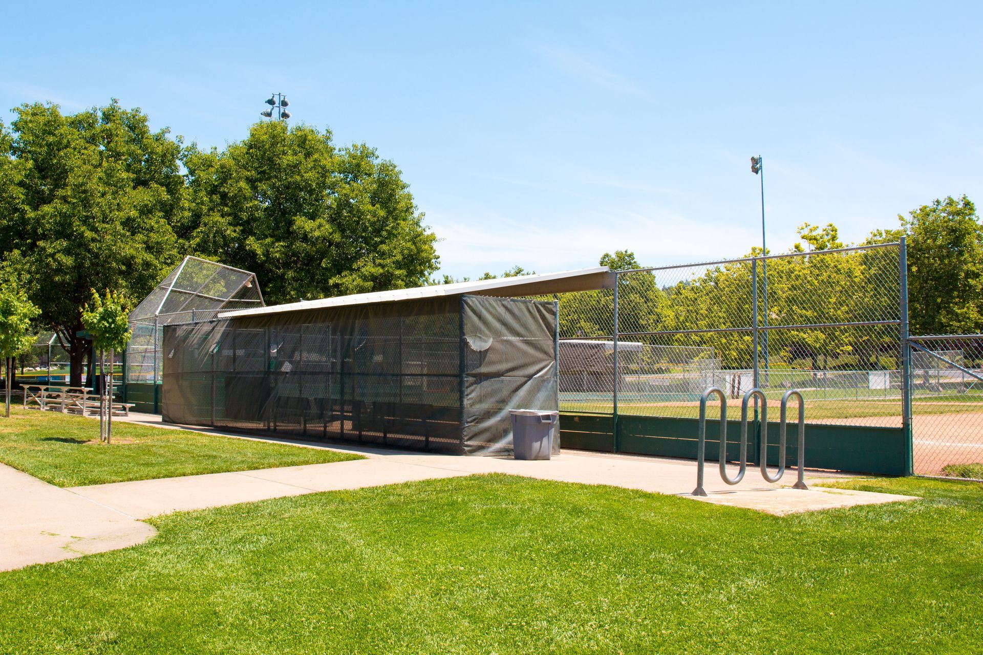 Outdoor bike shelter with a metal rack beside a grassy park path and chain-link fence.