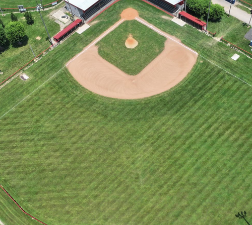 Aerial view of a baseball field with green outfield and tan infield, surrounded by grass and fences.