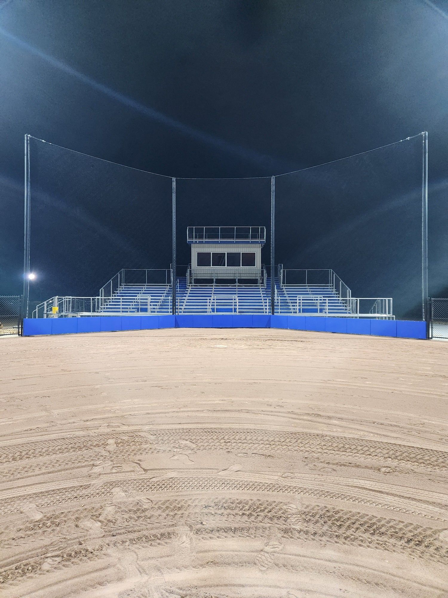 Empty nighttime rodeo arena with blue railings and a dirt floor under bright lights
