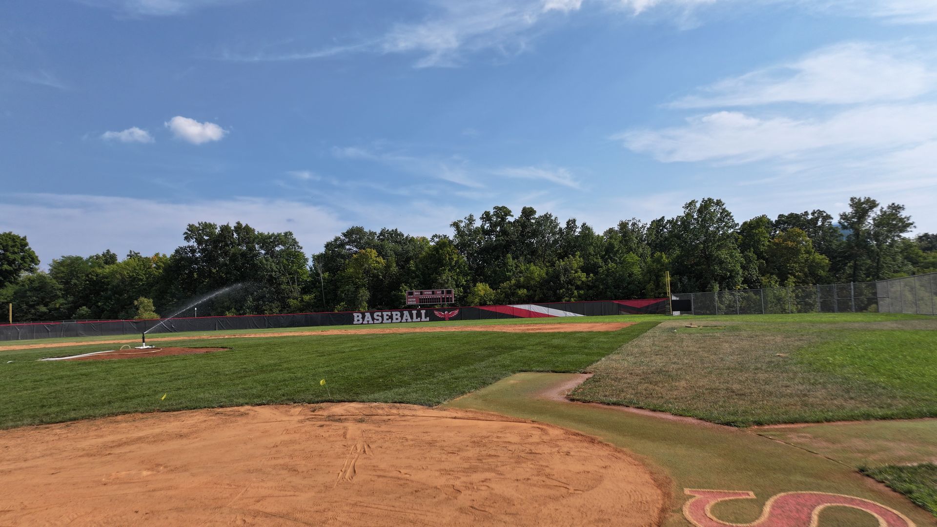 Baseball field with dirt infield, green outfield, and a few players in the distance under a blue sky.