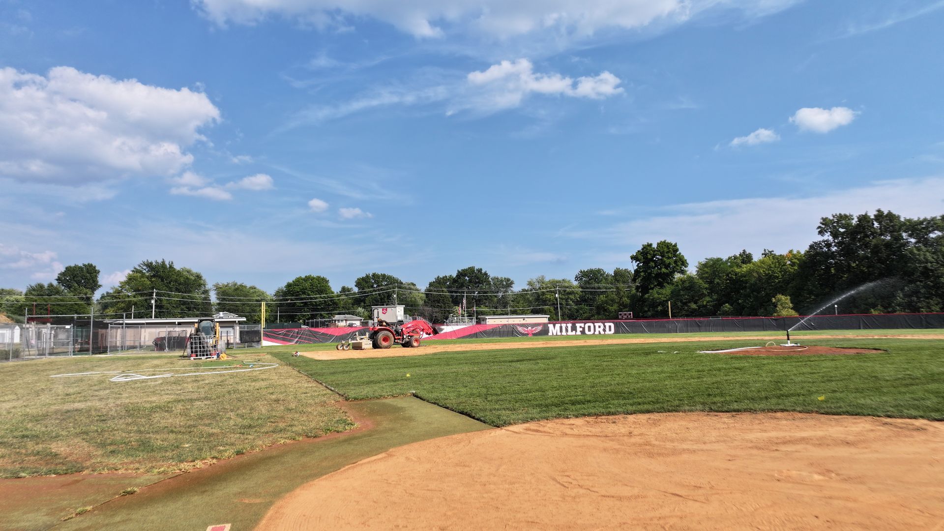 Baseball field under a blue sky, with players near the dugout and a sprinkler watering the outfield.