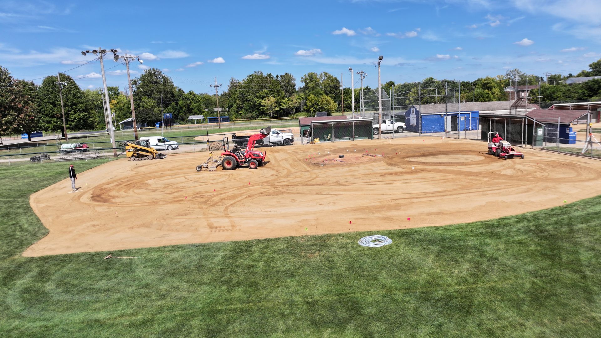 Construction vehicles grading a large dirt area beside a grassy park under a blue sky.