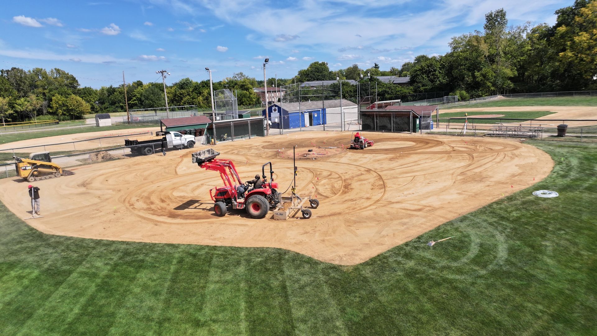 Red tractor grading a tan dirt infield beside a green sports field on a sunny day