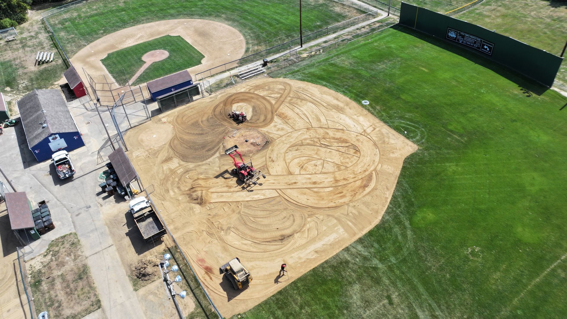 Aerial view of a dirt-covered baseball field with heavy machinery grading the infield beside green outfield grass.