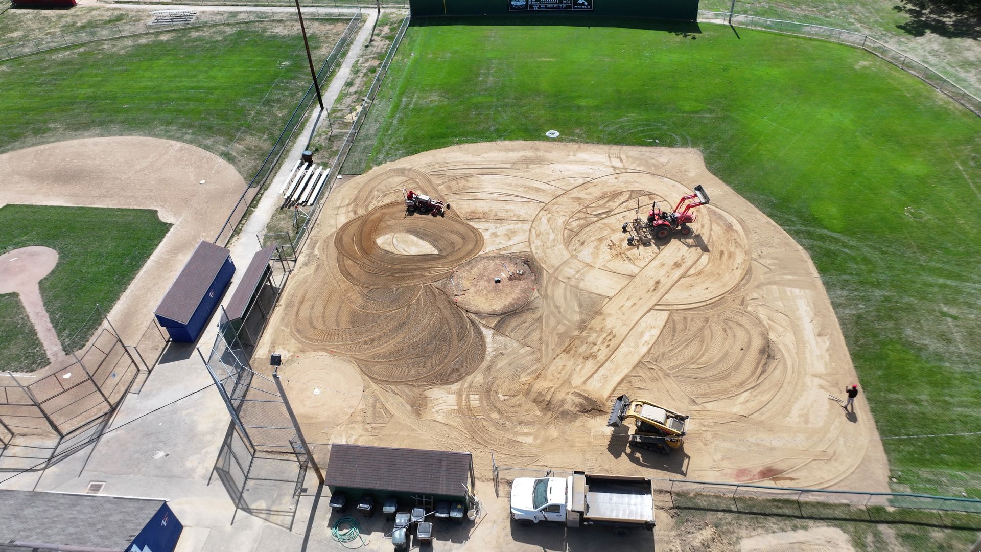 Aerial view of a dirt construction site with heavy machinery beside a grassy sports field.