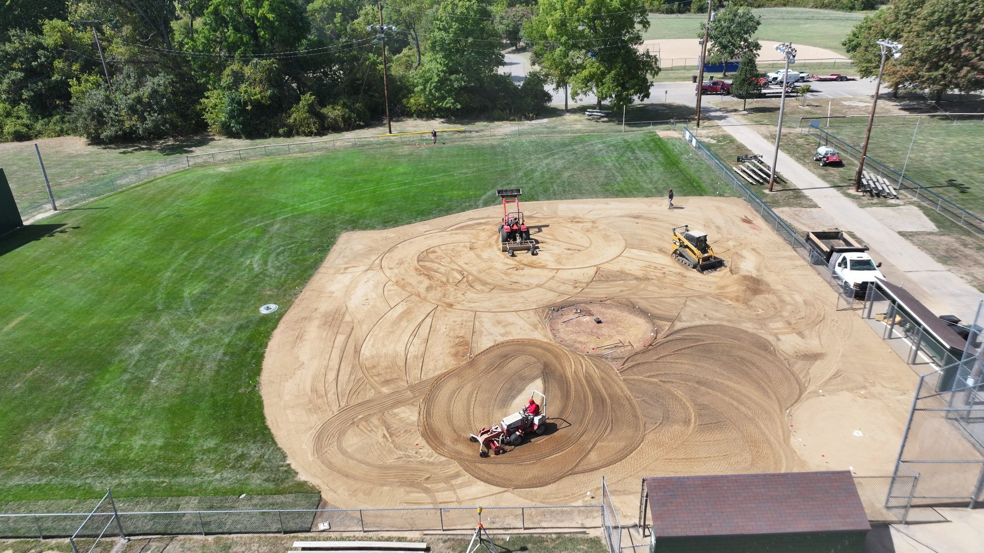 Aerial view of a dirt worksite with two excavators and a bulldozer leveling a large cleared area next to a grassy field.