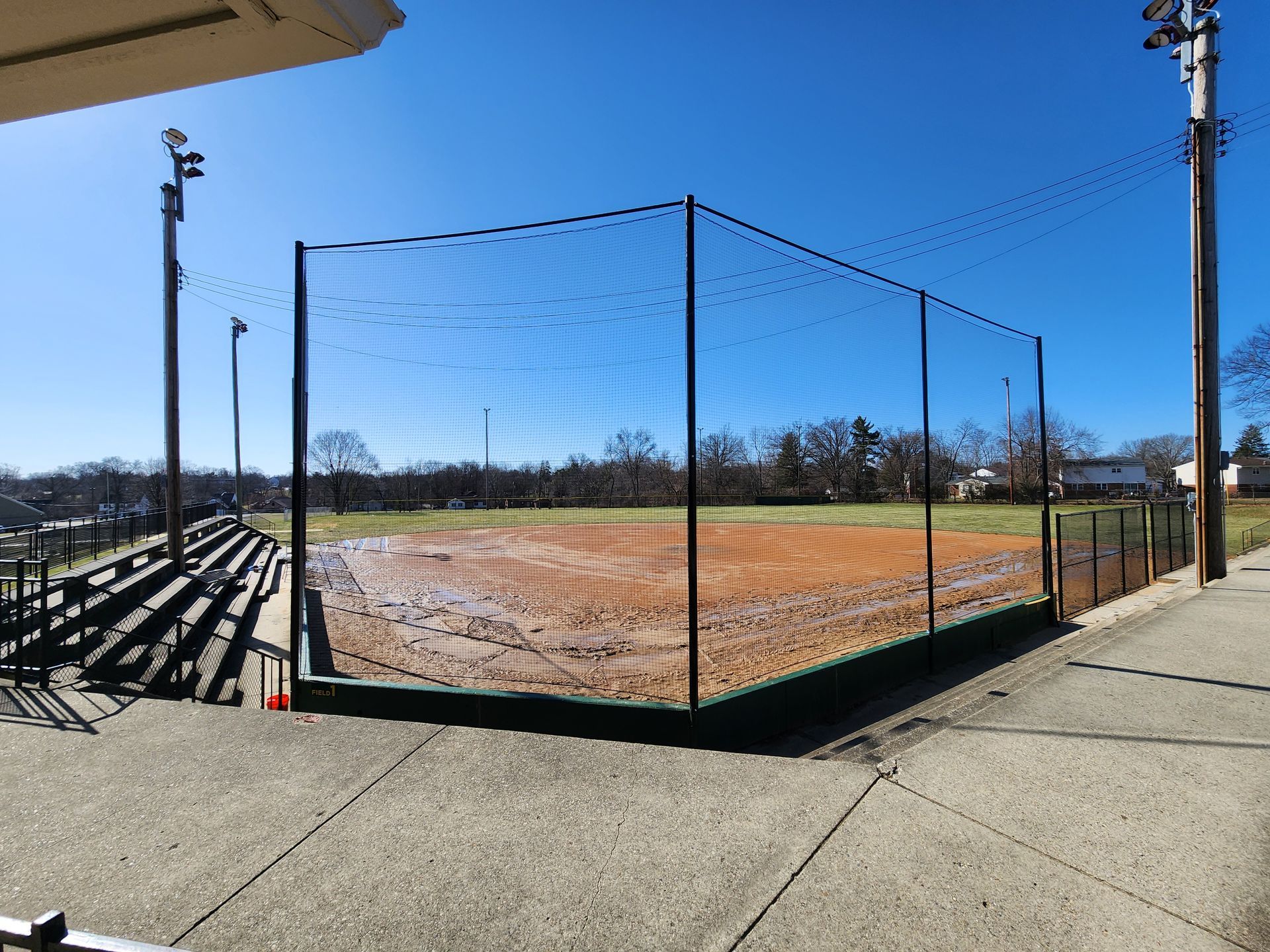 Empty fenced baseball field with muddy dirt, bordered by sidewalks under a clear blue sky.