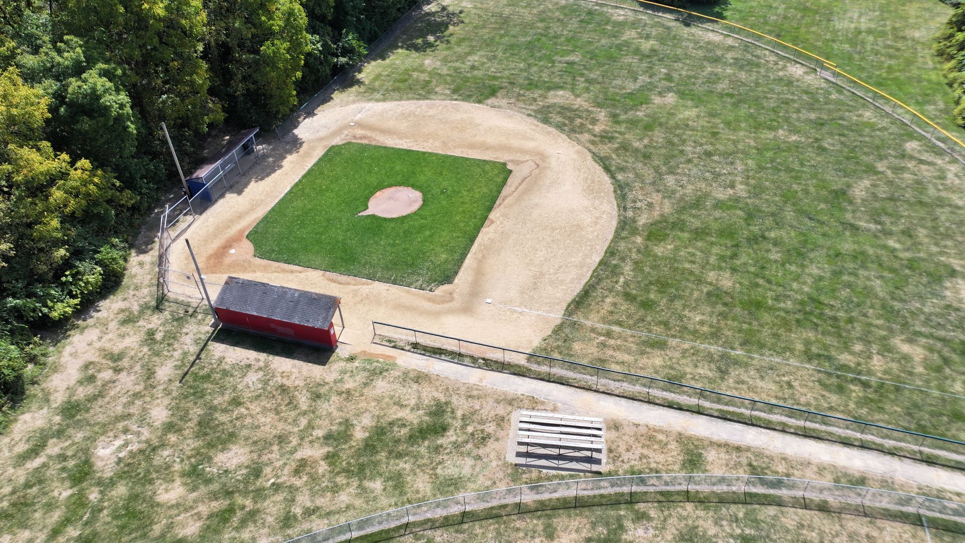 Aerial view of a small baseball field with a red dugout, bleachers, and surrounding grassy park.