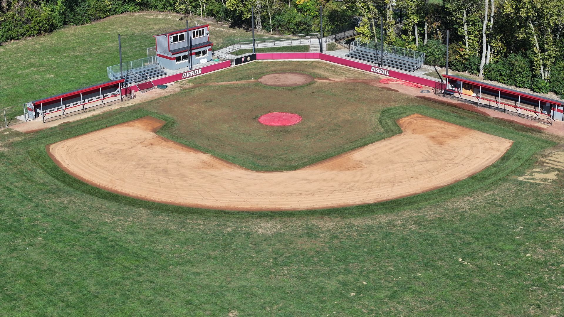 Aerial view of a small baseball field with red bleachers and a dirt infield surrounded by grass.